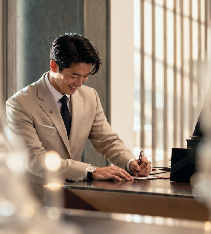 A hotel manager writes at a reception desk beside a bright window.