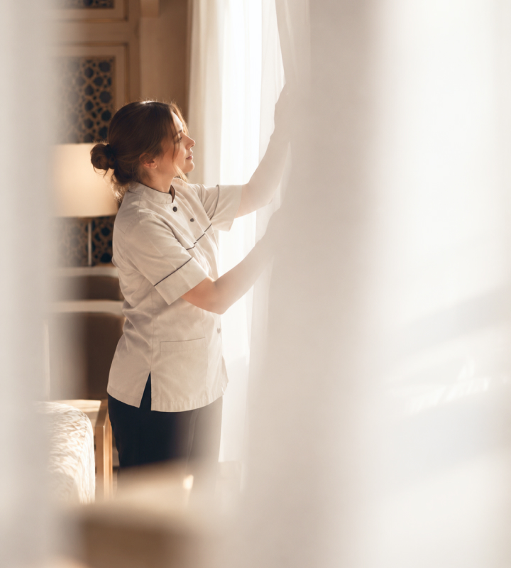 A hotel attendant opens sheer curtains in a sunlit guest room.